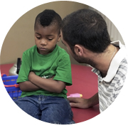 Adult speaking to young boy who is seated with his arms crossed.