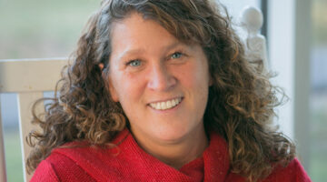 Jennifer Maeverde, a woman with long brown hair, blue eyes and a red knit sweater seated in a white wooden chair in an outdoor setting.