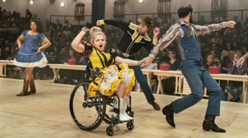 Actor Ali Stroker in a wheelchair pumping her fist with other actors dancing around her during Broadway revival of Oklahoma!