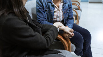 Two people seated next to each other in a waiting room holding hands.