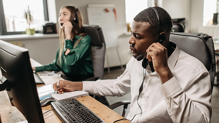 Two people, an Asian-American woman and an African-American man seated at desks in front of computers wearing headsets responding to calls.