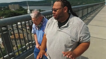 An African-American man and a senior white man with developmental disabilities walking along a pedestrian walkway overlooking a river.