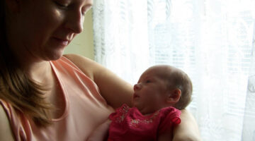 A young mother with long brown hair and a peach-colored top cradles her infant daughter in her left arm.