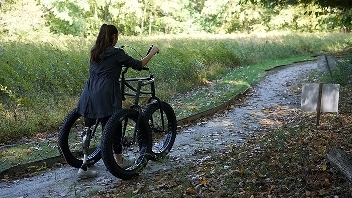 Adult woman with a prosthetic left leg walking outdoors on a dirt trail supported by a three-wheeled Afari "Off-Road" mobility device.