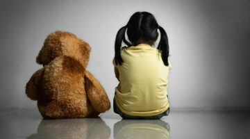 A stuffed brown bear and a young girl in a yellow shirt with black hair in pigtails sit with their backs to the camera.
