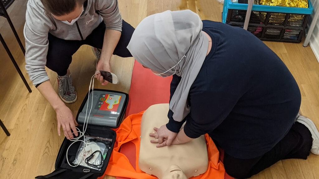 Female CPR instructor helping a woman wearing a hibaj kneeling on the floor doing chest compressions on a CPR training dummy.