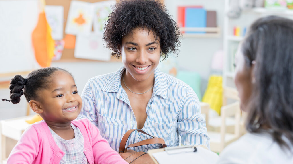 Young African American mom has a conference with her young daughter's preschool teacher. The little girl is sitting next to her mother.