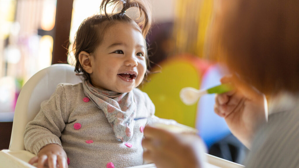 Smiling baby in a highchair being fed with a spoon by her mother.