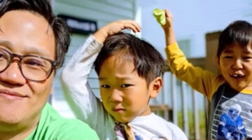 A selfie of a man of Asian descent with his two young sons standing behind him. One son looks annoyedand the other is laughing.