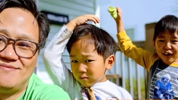 A selfie of a man of Asian descent with his two young sons standing behind him. One son looks annoyedand the other is laughing.