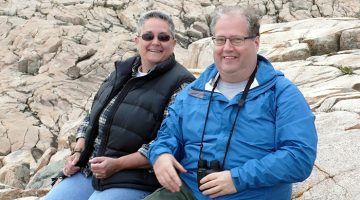A smiling middle aged woman with short graying hair, wearing glasses, a black vest and blue jeans, sits on large rocks with her brother, a smiling middle-aged man with thinning hair, who is wearing glasses, a blue rain jacket and khaki pants with a pair of binoculars hanging from his neck.