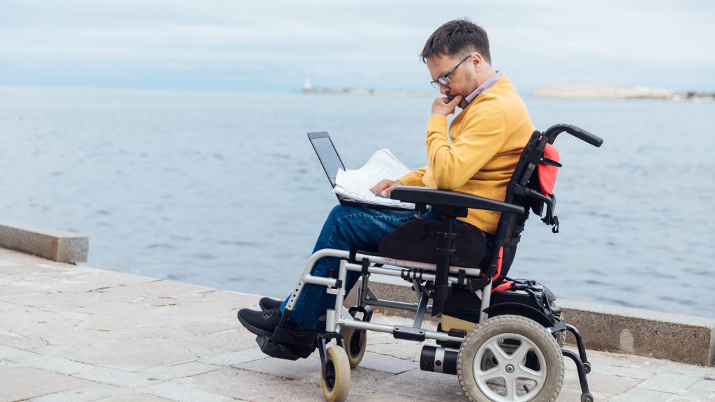 Maine sitting in a wheelchair with a laptop on his lap.
