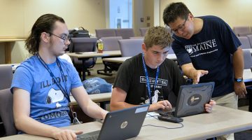 Two students seated in a college lecture hall using their computers to interact with a ChatGPT model as a professor looks on.