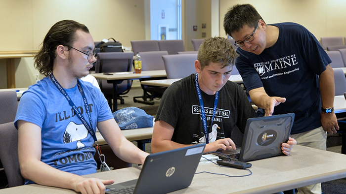 Two students seated in a college lecture hall using their computers to interact with a ChatGPT model as a professor looks on.