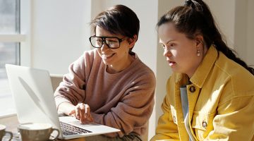 Two women seated next to each other, looking at a computer screen. The woman on the left has short brown hair, glasses and a pink sweater. The woman on the right has Down syndrome, long brown hair and is wearing a yellow denim jacket.