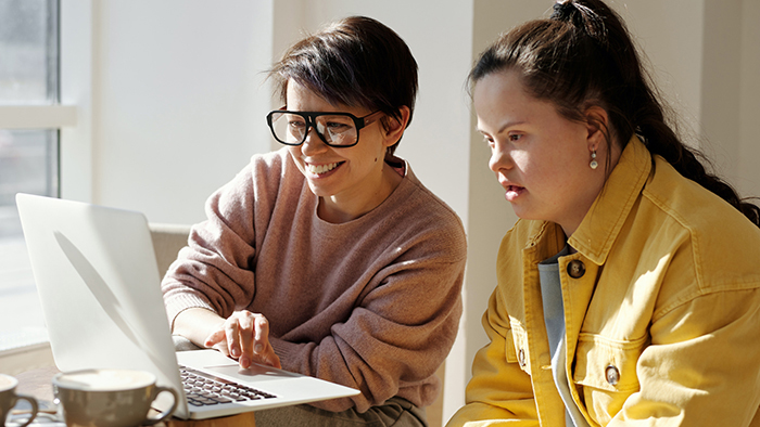 Two women seated next to each other, looking at a computer screen. The woman on the left has short brown hair, glasses and a pink sweater. The woman on the right has Down syndrome, long brown hair and is wearing a yellow denim jacket.