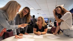 Four women standing around a round table facing each other, engaged in a discussion.