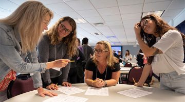Four women standing around a round table facing each other, engaged in a discussion.