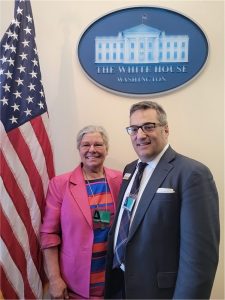 Two people, a woman and a man, standing in the White House complex next to an indoor American flag, with the White House shield above their heads.