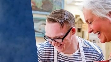 Smiling young adult with Down syndrome standing next to a coworker in a restaurant setting, clearing a table of cups.